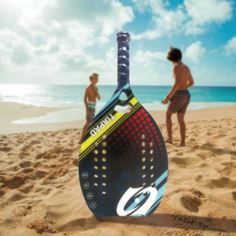 Raquete de padel Tapard colorida apoiada na areia da praia, com o mar e turistas ao fundo em cenário de lazer esportivo.
