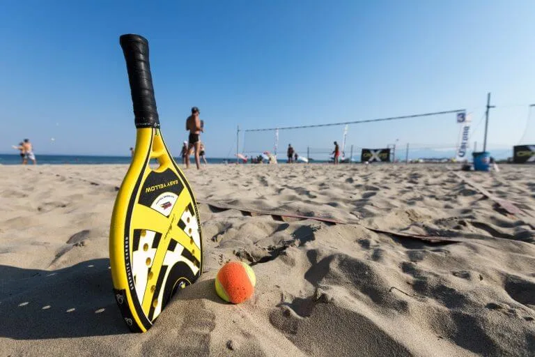Raquete de beach tennis Gravity Flow amarela e preta na areia da praia com bola colorida, quadra e jogadores ao fundo em cenário ensolarado.