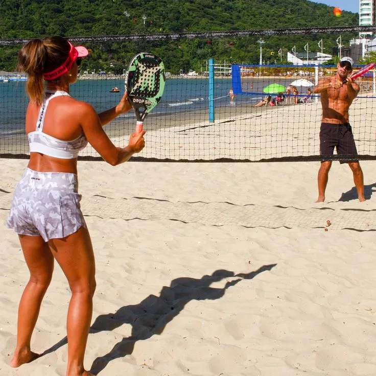 Dois jogadores de beach tennis em quadra de areia à beira-mar, com raquetes e bola laranja em jogo, cenário de praia com colinas e pessoas ao fundo.