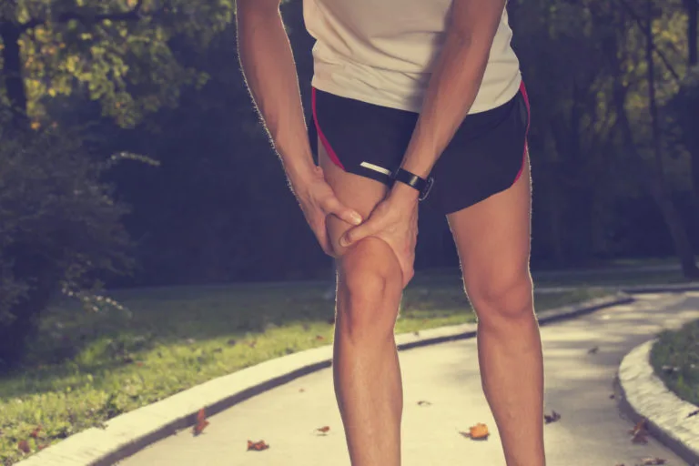Pessoa em trilha ao ar livre, vestindo camiseta branca e shorts pretos, segurando o joelho direito com as mãos em sinal de dor ou desconforto.