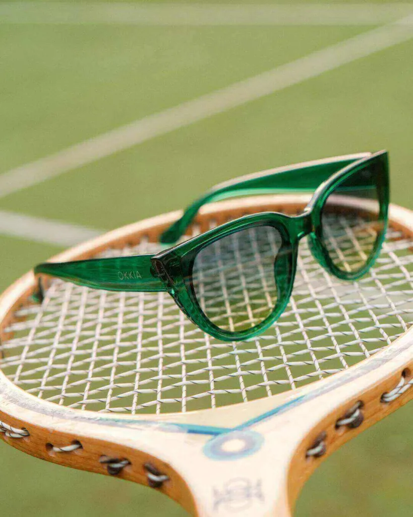 Óculos esportivos verdes translúcidos OKKIA apoiados em raquete de tênis vintage em quadra verde.