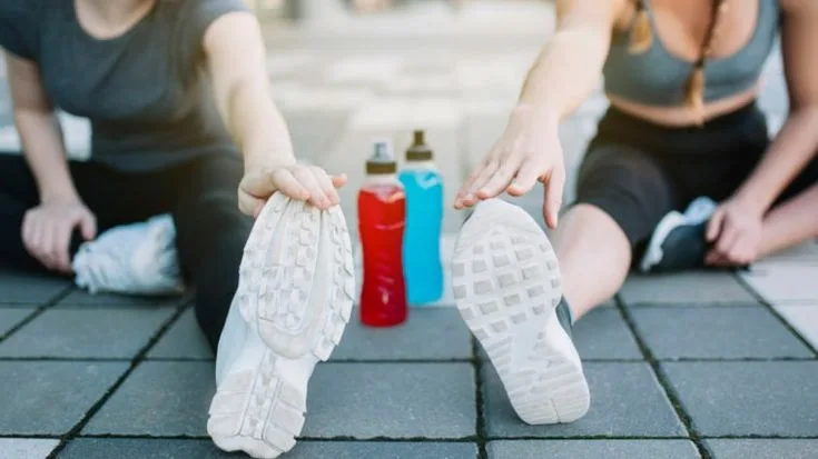 Dois atletas alongando pernas em área externa, com garrafas de bebida esportiva vermelha e azul entre eles.