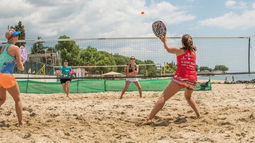 Quatro jogadores de beach tennis em quadra de areia à beira-mar, com raquetes e bola laranja em jogo, destacando ação em dupla e trabalho em equipe.