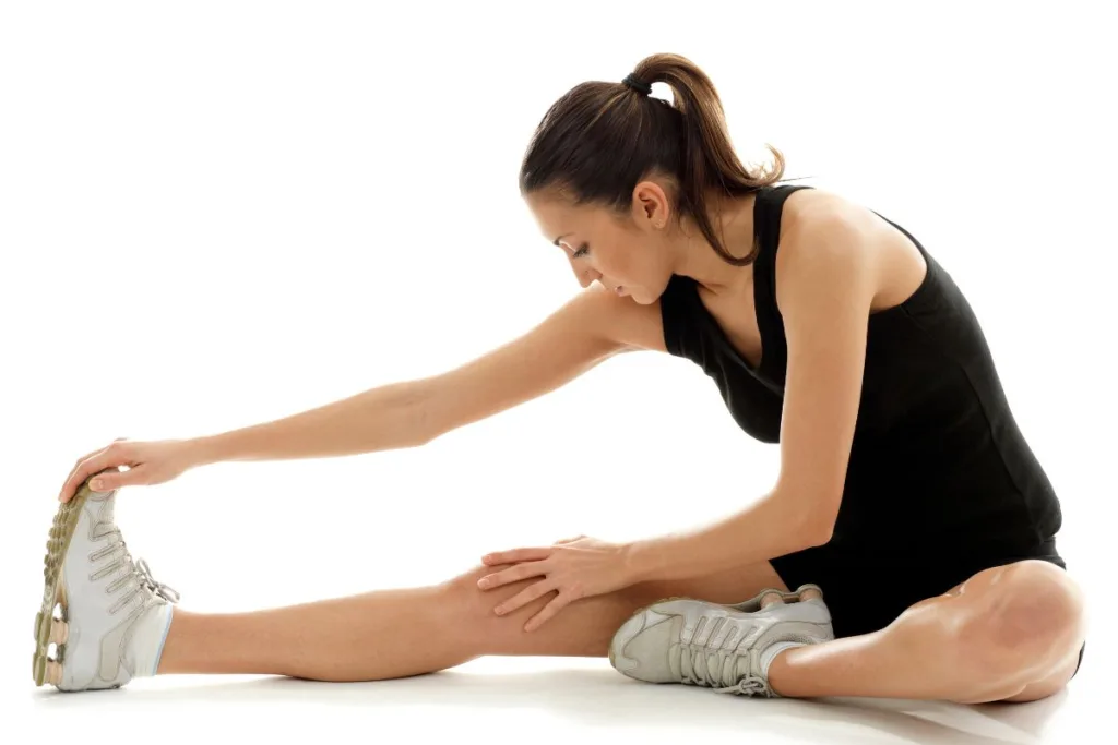 Pessoa sentada no chão realizando alongamento de perna, com uma perna estendida e a outra dobrada, tocando os dedos do pé para trabalhar flexibilidade e recuperação muscular.
