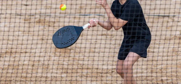 Jogador de beach tennis em quadra de areia, vestindo camiseta preta e shorts pretos, preparando golpe com raquete contra bola amarela e vermelha.