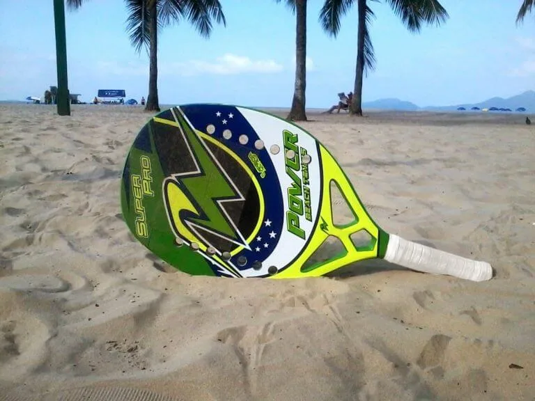 Raquete de beach tennis colorida fincada na areia de praia tropical com palmeiras ao fundo.