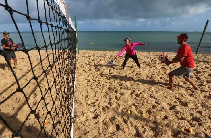 Três pessoas jogando beach tennis em quadra de areia à beira-mar, com bolas espalhadas e oceano ao fundo.