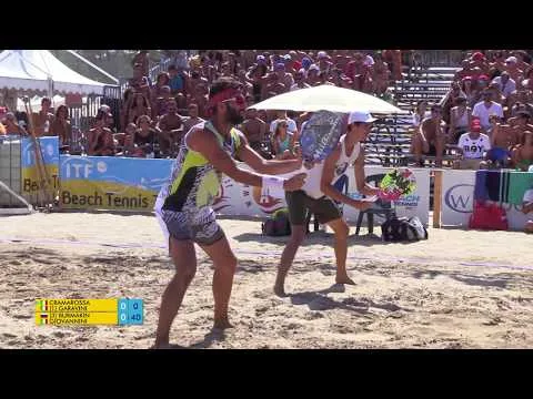 Jogadores de beach tennis em quadra de areia durante partida profissional, com torcida em arquibancadas ao fundo.