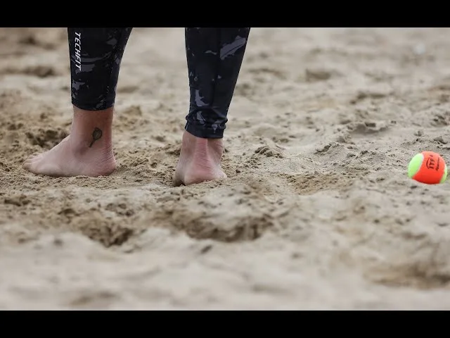 Pessoa descalça em areia com calça legging escura e bola de tênis amarela e laranja ao lado.