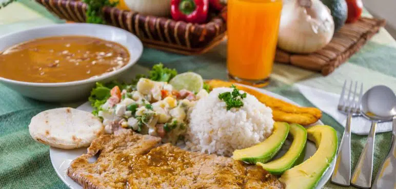 Prato latino-americano com carne grelhada, arroz branco, abacate, banana frita, arepa, salada de legumes, sopa e suco de laranja.
