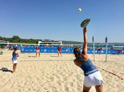 Jogadoras de beach tennis em quadra de areia, atleta da Bulgária realizando saque contra adversárias de uniforme vermelho.