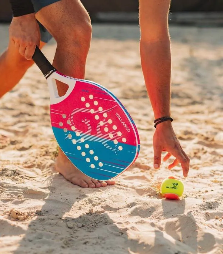 Jogador de beach tennis descalço em quadra de areia, segurando raquete colorida da marca Kallango e bola amarela.