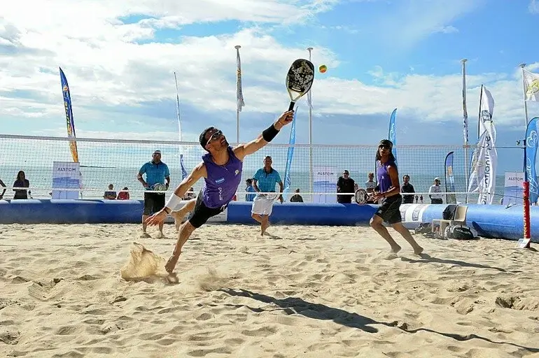 Jogador de beach tennis mergulhando na areia para devolver bola, em partida competitiva com torcida e banners ao fundo.