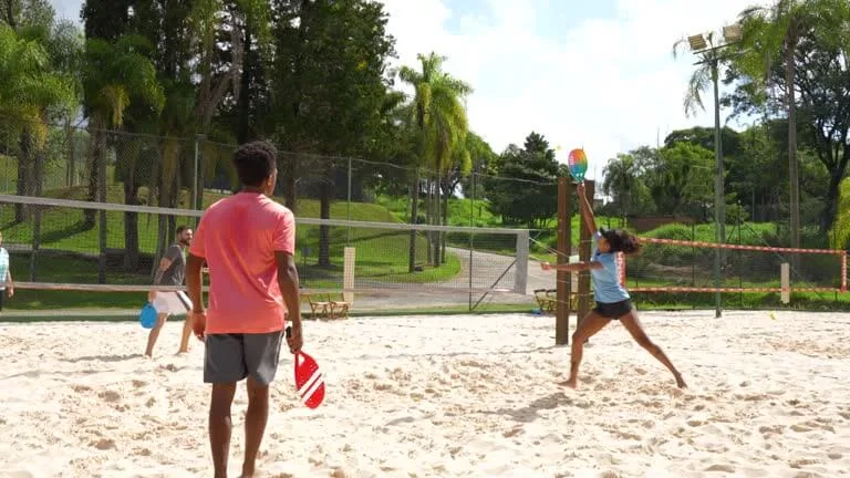 Jogadores de beach tennis em quadra de areia cercada por palmeiras, realizando jogada intensa sob céu ensolarado.