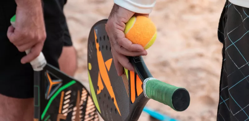 Dois jogadores segurando raquetes de beach tennis com detalhes coloridos e bola amarela em quadra de areia.