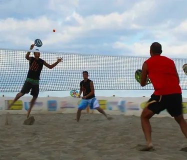 Quatro jogadores disputando partida de beach tennis em quadra de areia, com bola no ar acima da rede.