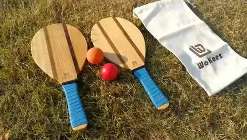 Conjunto de frescobol com duas raquetes de madeira de cabo azul, duas bolas coloridas e bolsa de transporte branca personalizada.
