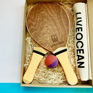 Conjunto de frescobol com duas raquetes de madeira, bola e toalha personalizada, organizados em caixa ecológica.