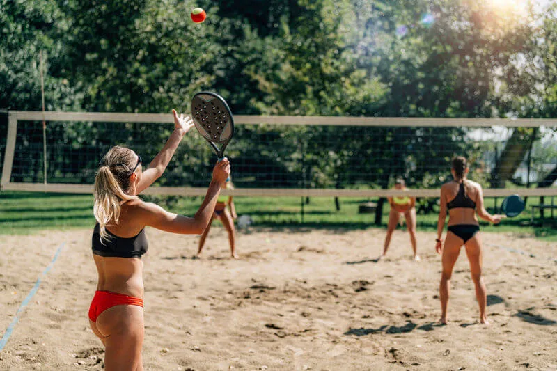 Jogadores praticando beach tennis em quadra de areia ao ar livre, atleta preparando saque com raquete e bola no ar.