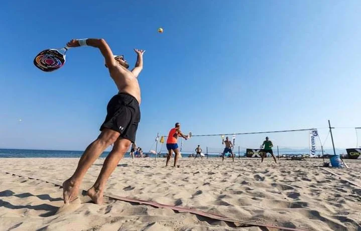 Jogadores de beach tennis em praia ensolarada, atleta saltando para sacar bola sobre a rede.