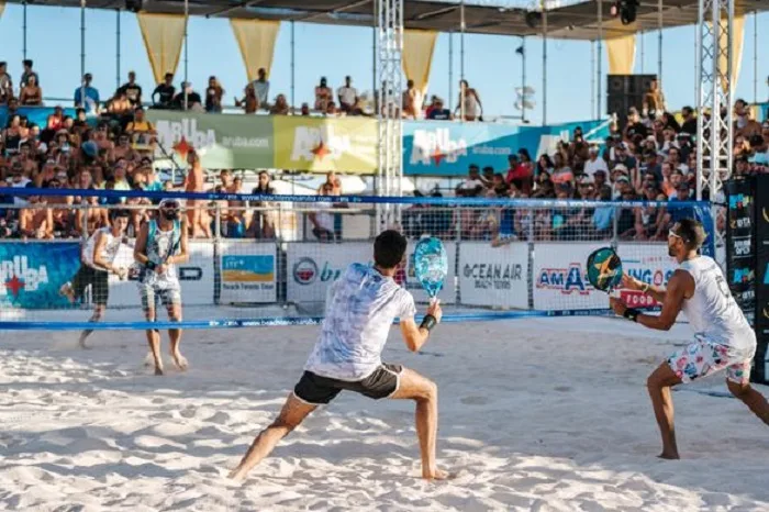 Partida de beach tennis em quadra de areia, quatro jogadores em ação diante de público numeroso sob sol intenso.