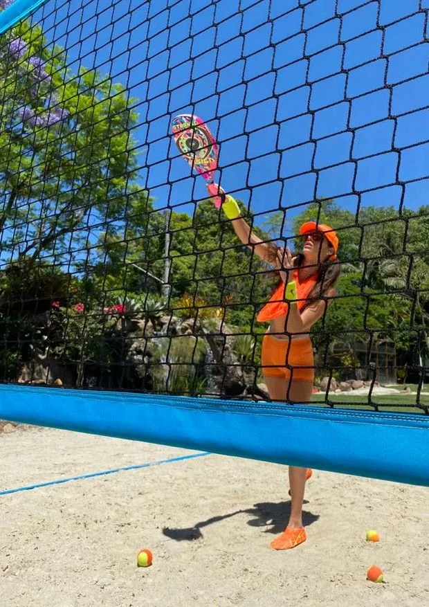 Jogador de beach tennis em quadra de areia, vestido com uniforme laranja e boné, realizando golpe com raquete colorida contra bola, em cenário ensolarado com árvores ao fundo.