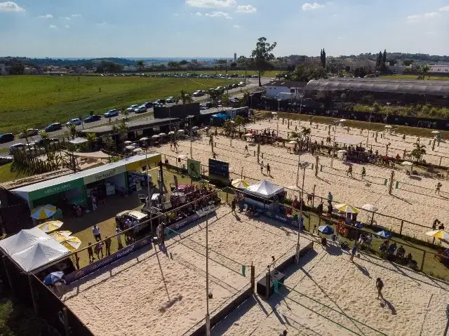 Vista aérea de complexo esportivo com várias quadras de areia para vôlei, jogadores em ação e tendas de patrocinadores ao redor.
