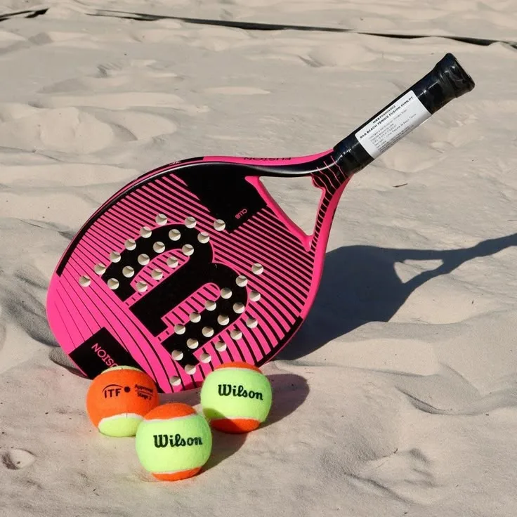 Raquete de padel Wilson rosa sobre areia com quatro bolas de tênis, ideal para beach tennis e padel.