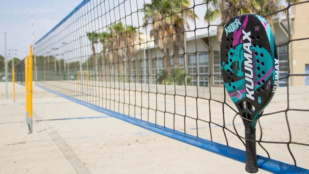 Raquete colorida de beach tennis pendurada em rede de quadra de areia, com palmeiras e prédio ao fundo em dia ensolarado.