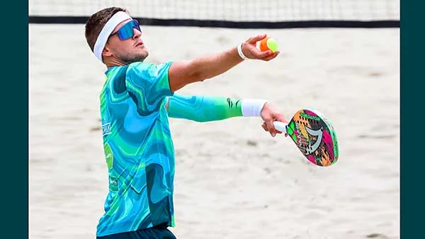 Jogador de beach tennis em quadra de areia, usando camiseta azul e verde estampada, faixa branca e óculos esportivos, preparando saque com raquete colorida contra bola amarela e laranja.