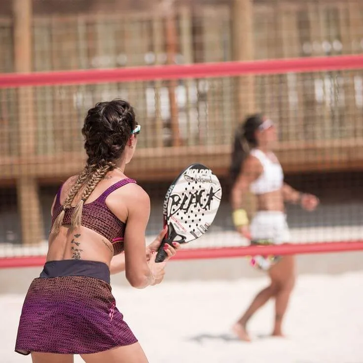 Jogadores de beach tennis em quadra de areia, usando roupas esportivas coloridas e raquetes RUNK.