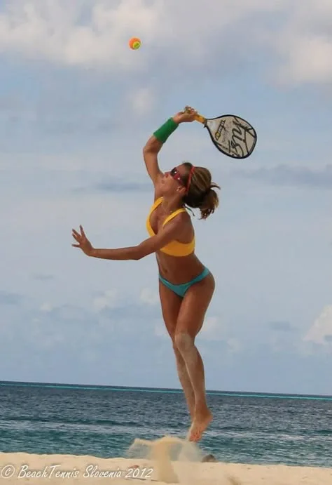 Jogador de beach tennis saltando na areia para golpear bola, com mar ao fundo e céu parcialmente nublado.