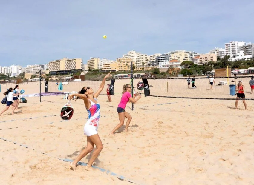 Grupo de pessoas jogando beach tennis em quadra de areia na praia, com prédios ao fundo em dia ensolarado.
