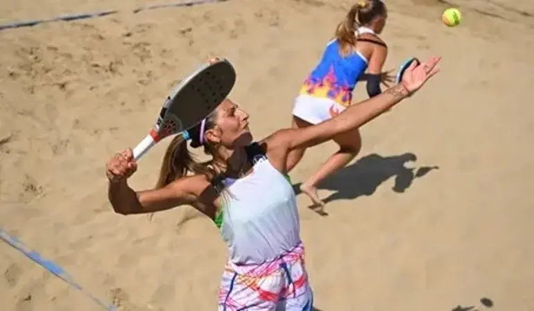 Jogador de beach tennis preparando saque em quadra de areia, com raquete na mão e bola lançada ao ar, enquanto adversário aguarda a jogada.