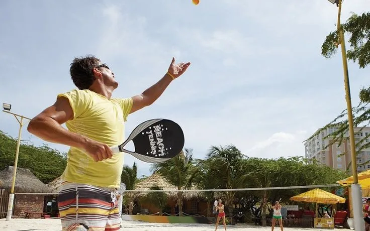Jogador de beach tennis em quadra de areia tropical, vestindo camiseta amarela e shorts listrados, prestes a golpear bola laranja com raquete.