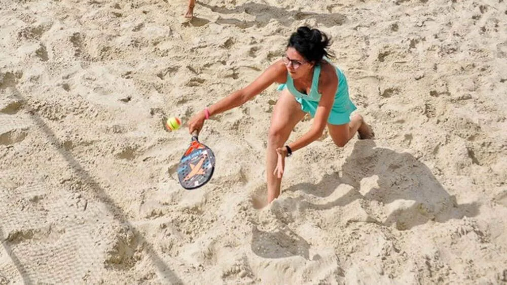 Jogador de beach tennis em quadra de areia, vestindo roupa esportiva turquesa e óculos escuros, realizando voleio com raquete contra bola amarela e vermelha.