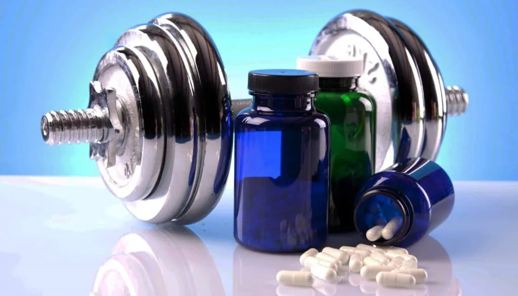 Dois halteres cromados sobre superfície refletiva ao lado de frascos de suplementos esportivos e cápsulas brancas espalhadas.