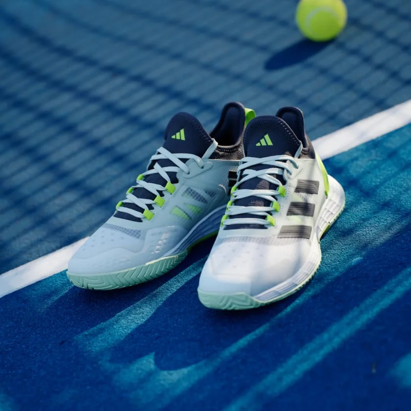 Par de tênis Adidas brancos com detalhes verde neon e azul marinho sobre quadra de tênis azul, acompanhado de bola amarela.