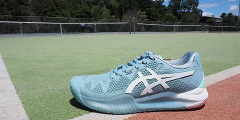 Tênis Asics azul claro com detalhes brancos sobre quadra de tênis verde e vermelha, próximo à rede.