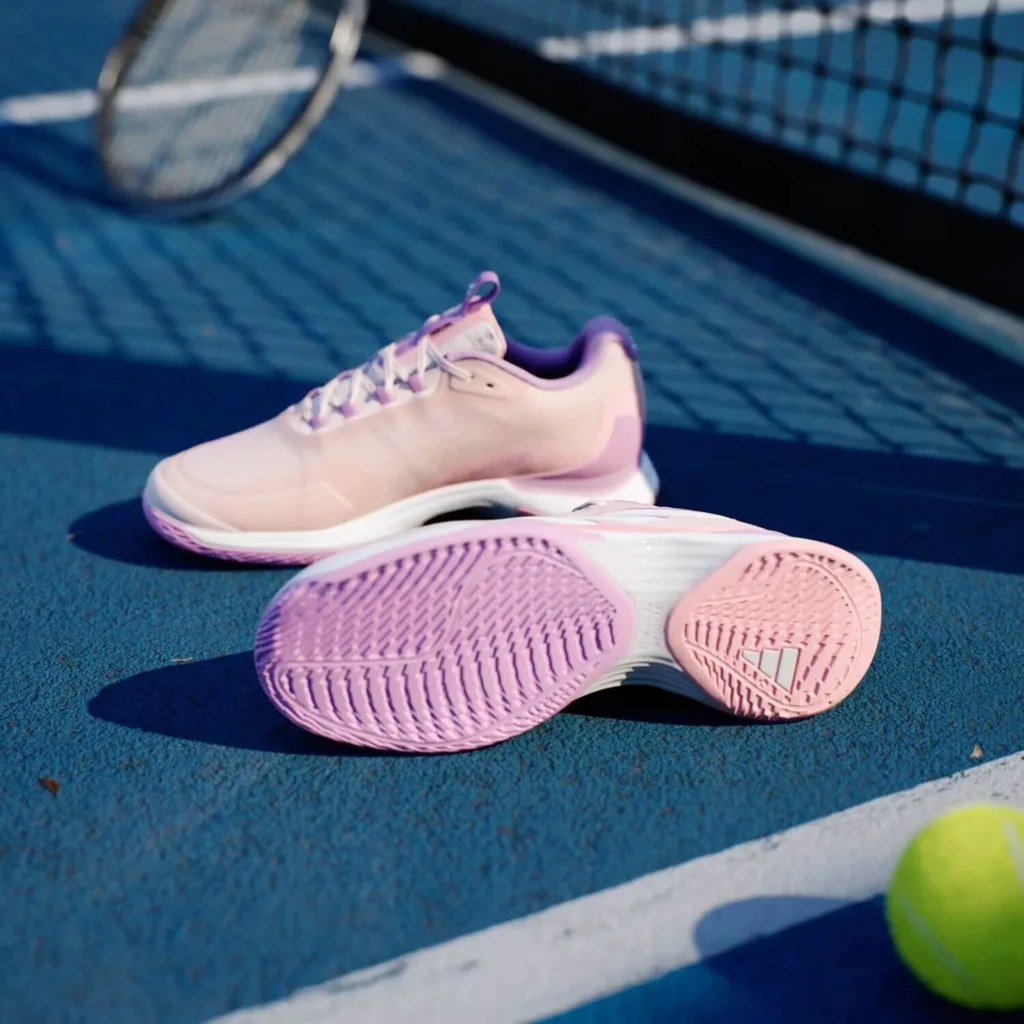 Tênis esportivos rosa e roxo da linha beach tennis posicionados em quadra azul, próximos a bola e raquete.