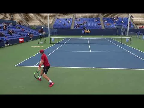 Partida de tênis em quadra dura da UCLA com dois jogadores em ação, um preparando golpe de fundo e outro na defesa.