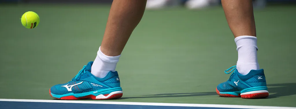 Tênis esportivos Mizuno azul turquesa com solado vermelho em quadra de tênis verde, próximos a bola amarela em movimento.