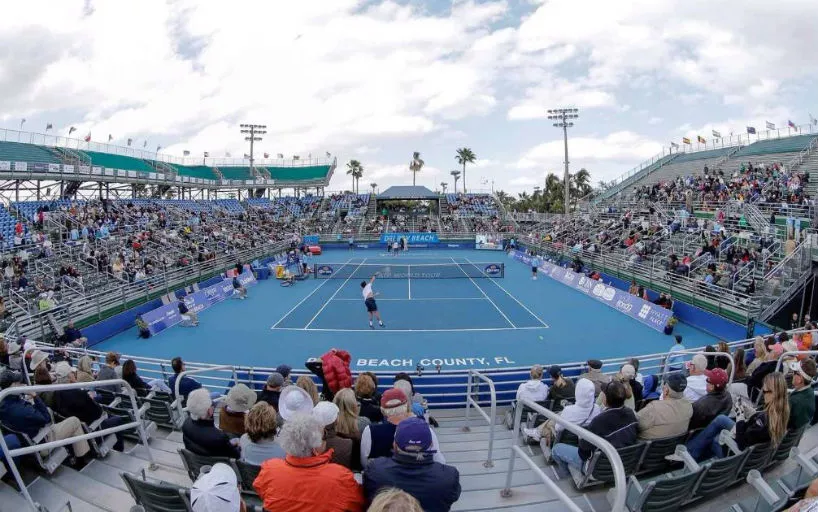 Partida profissional de tênis em quadra dura azul, com jogadores em ação e arquibancadas cheias em Palm Beach County, Flórida.