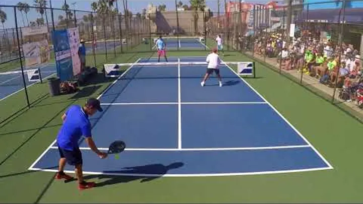 Jogadores de pickleball em quadra externa azul e verde, atleta de camiseta azul preparando saque diante de torcida.