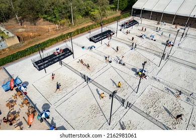 Vista aérea de complexo esportivo com várias quadras de vôlei de areia, jogadores em ação e áreas de descanso com guarda-sóis.