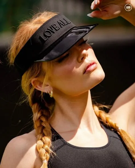 Pessoa com cabelo trançado claro usando viseira preta com borda brilhante e inscrição “LOVE ALL”, levantando a mão para proteger os olhos da luz.