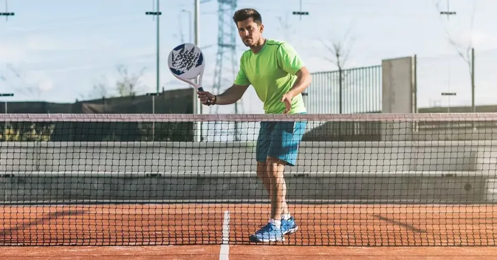 Homem jogando padel em quadra de saibro, vestindo camiseta verde neon, shorts azuis e tênis esportivos azuis, segurando raquete branca com detalhes azuis próximo à rede, em posição de ataque.