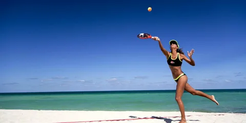 Mulher jogando beach tennis na areia branca de uma praia tropical, vestindo top e shorts pretos com detalhes neon, raquete levantada para rebater bola amarela sob céu azul intenso e mar verde-turquesa ao fundo.