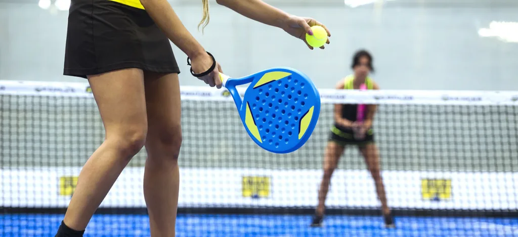 Jogadora de padel em quadra azul, preparando o saque com bola amarela na mão e raquete azul com detalhes amarelos, enquanto adversária aguarda próxima à rede.