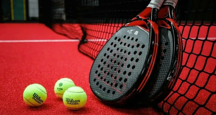 Duas raquetes de padel Wilson com design preto e vermelho apoiadas na rede de uma quadra de piso vermelho, acompanhadas por três bolas Wilson Padel amarelas posicionadas próximas à base das raquetes.